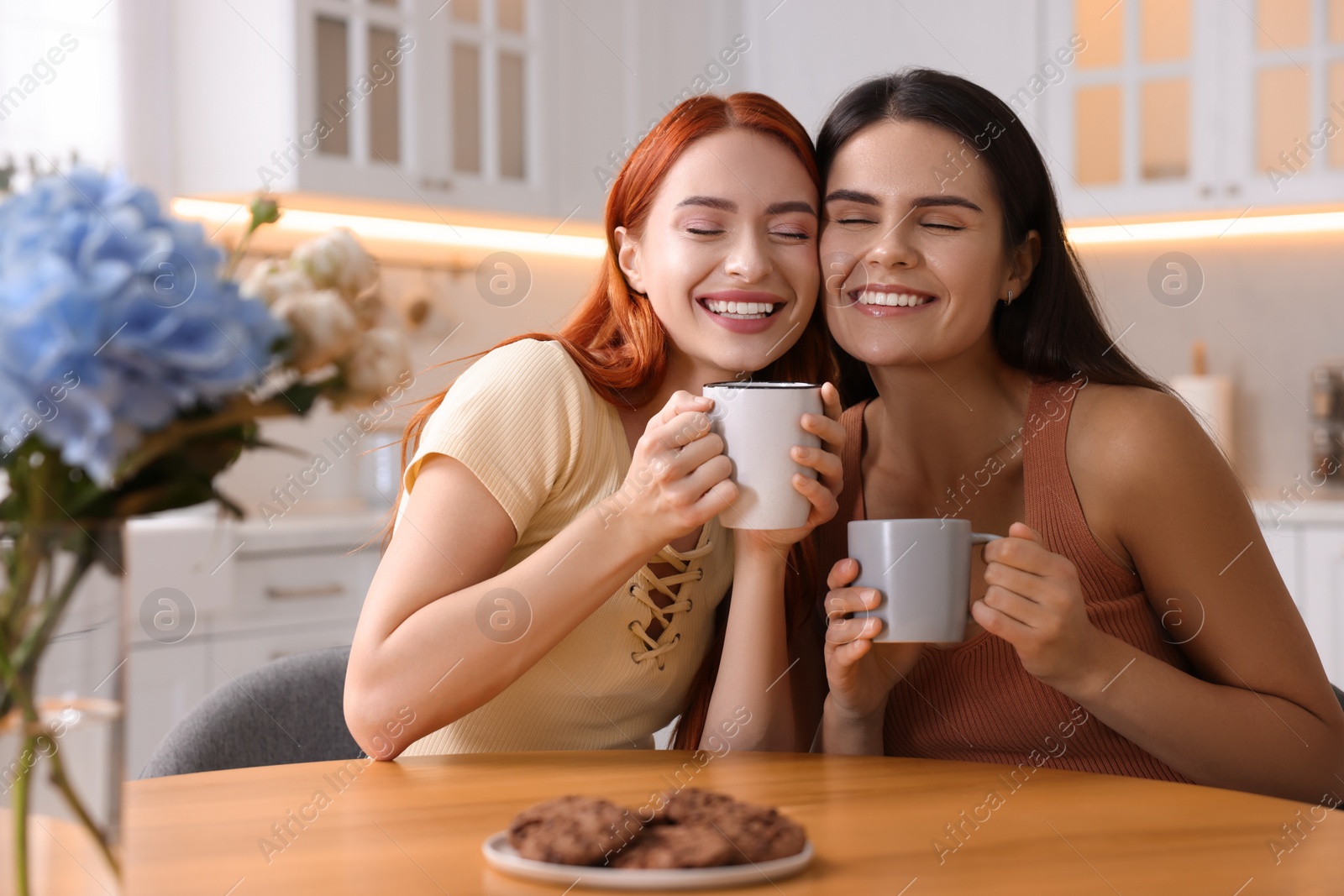 Photo of Happy young friends with cups of drink spending time together at table in kitchen