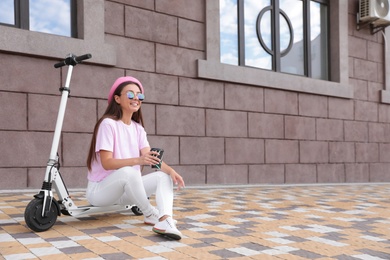 Photo of Young woman with cup of coffee sitting on electric kick scooter outdoors