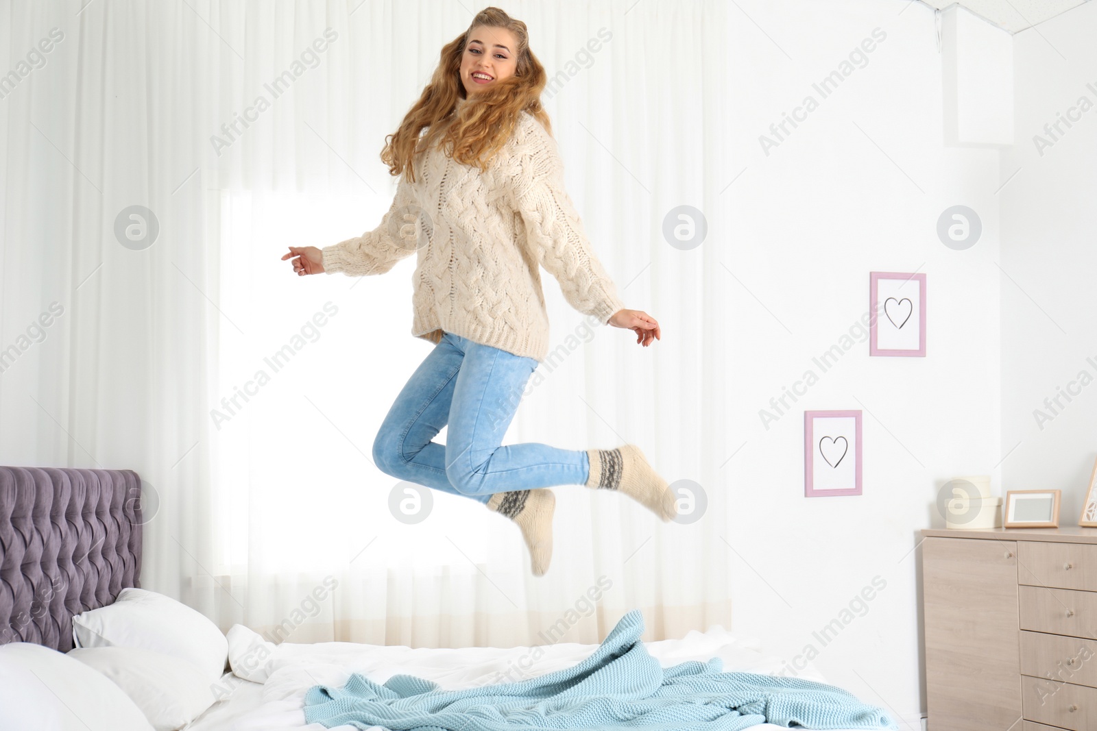 Photo of Attractive smiling young woman in cozy warm sweater jumping on bed at home