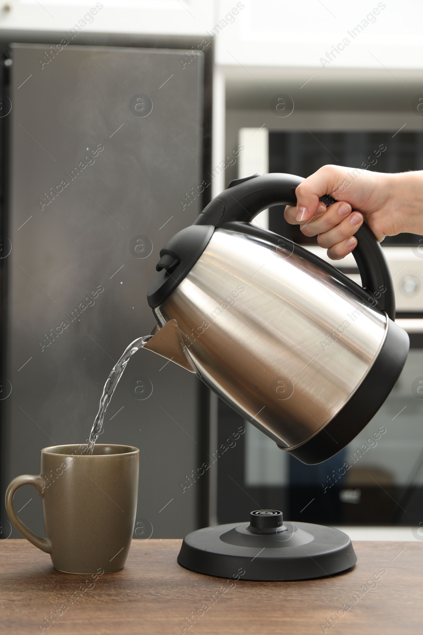 Photo of Woman pouring hot water from electric kettle into cup in kitchen, closeup