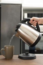 Photo of Woman pouring hot water from electric kettle into cup in kitchen, closeup