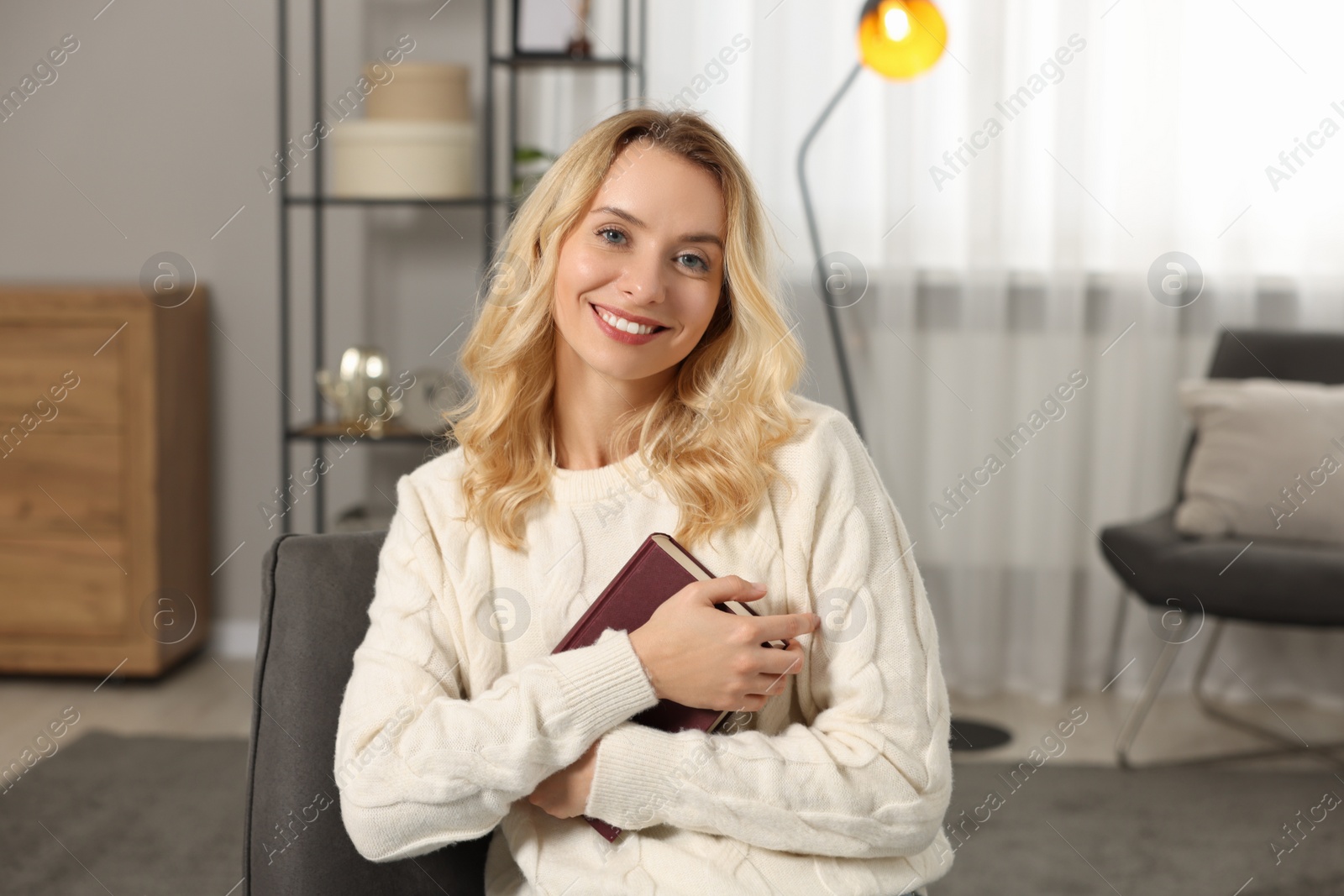 Photo of Happy woman in stylish warm sweater holding book at home