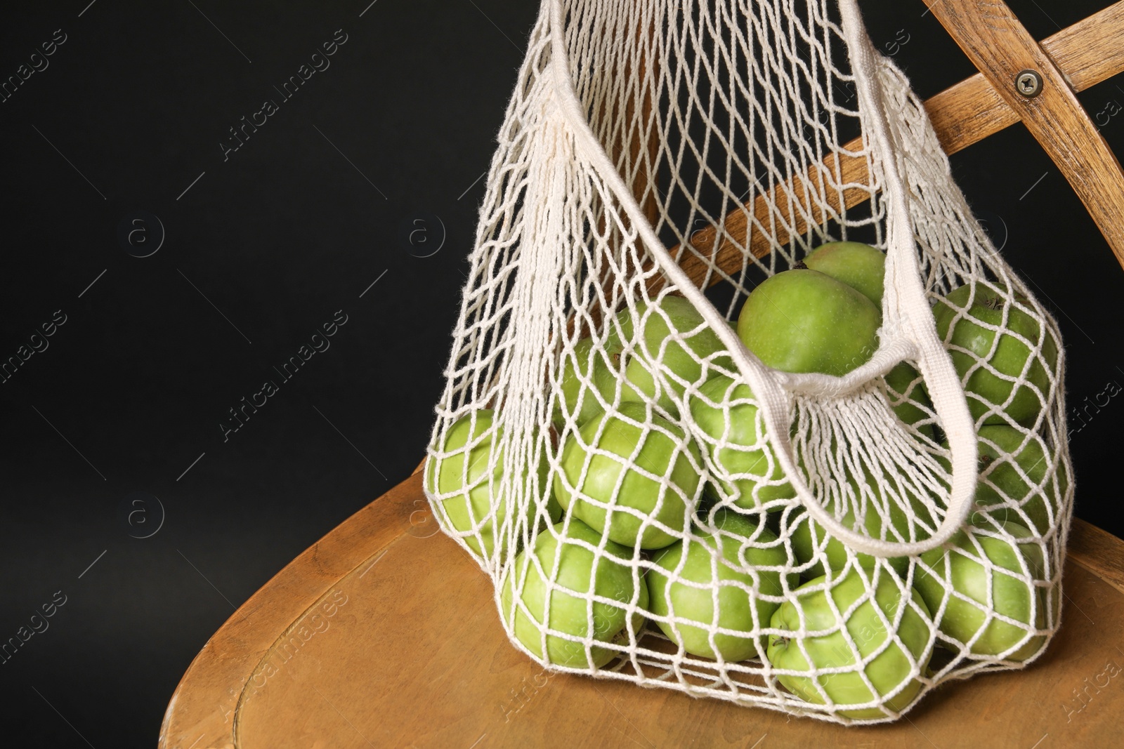 Photo of Green apples in net bag on wooden chair against black background. Space for text