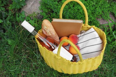 Yellow wicker bag with book, peaches, baguette and wine on green grass outdoors, above view