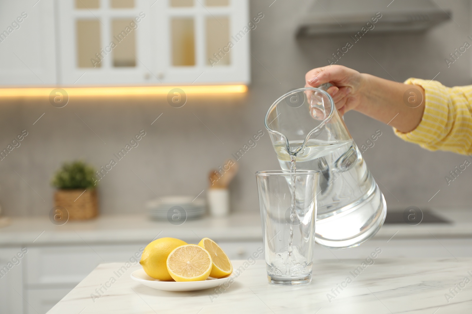 Photo of Woman pouring water from jug into glass at white table in kitchen, closeup. Space for text