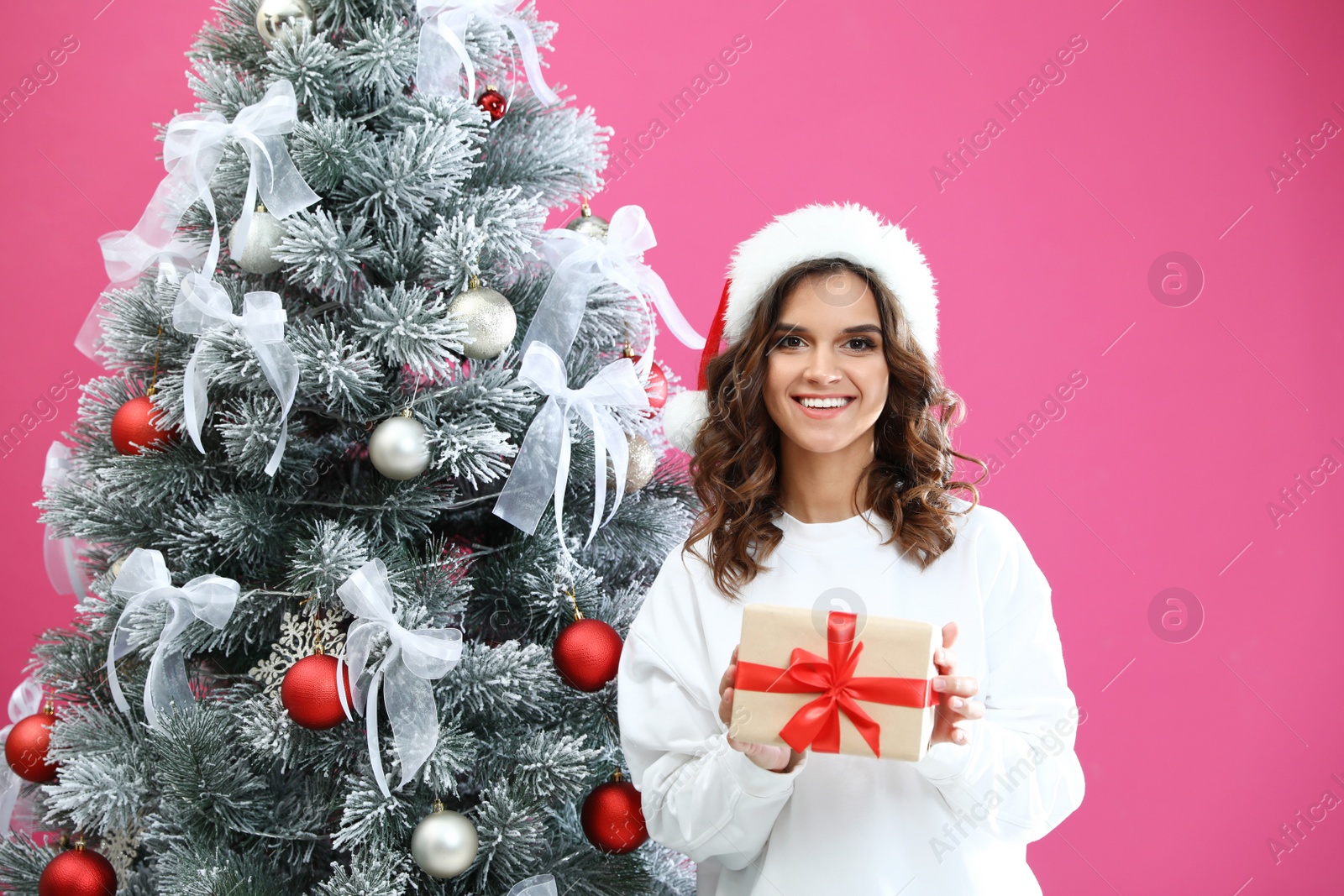 Image of Happy young woman in Santa hat with gift near Christmas tree on pink background