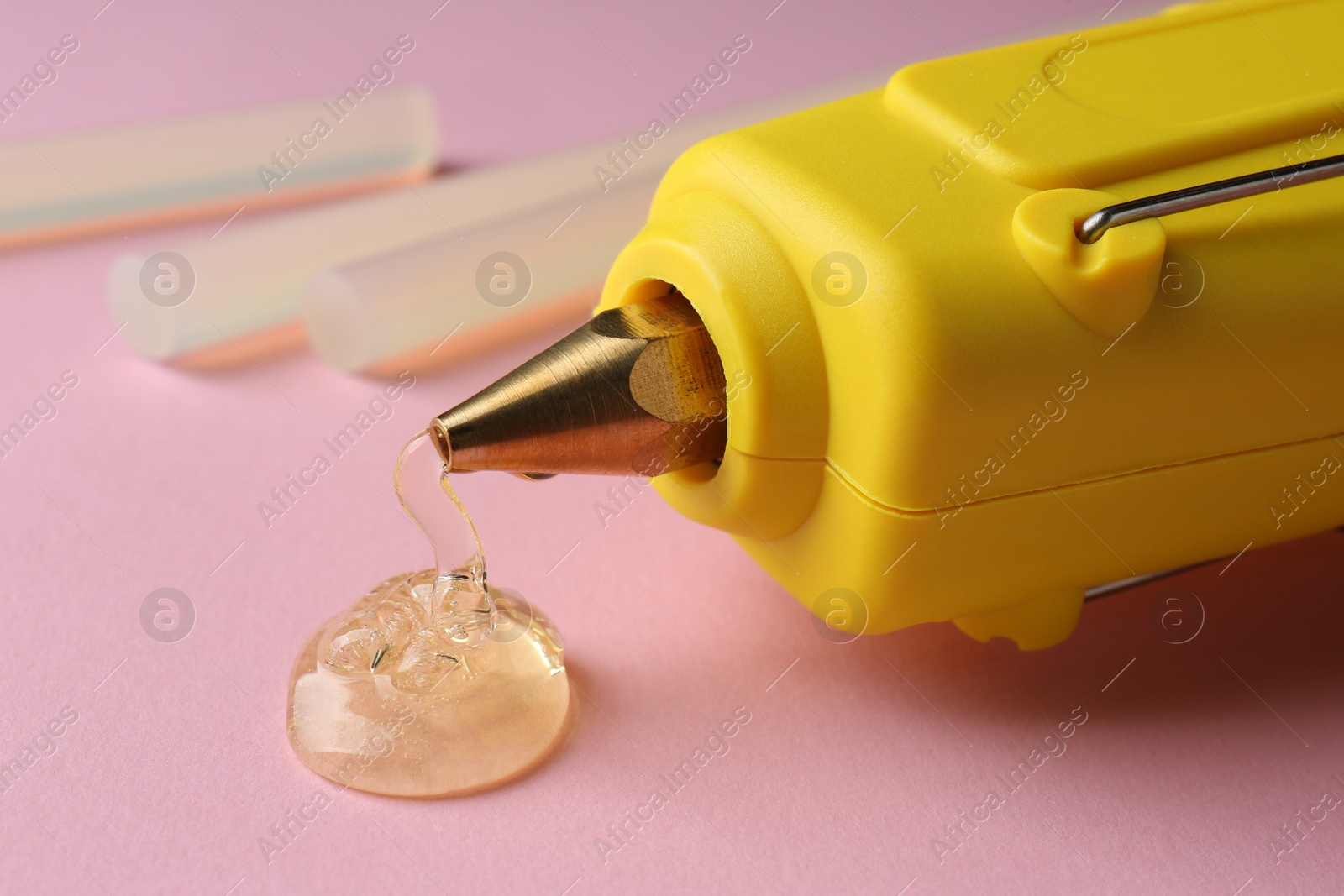 Photo of Melted glue dripping out of hot gun nozzle near sticks on pink background, closeup