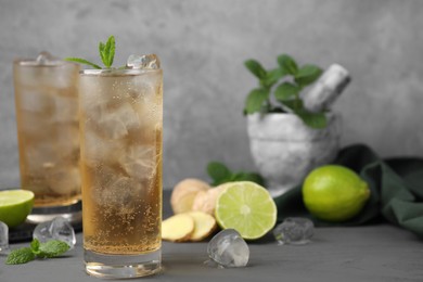 Photo of Glass of tasty ginger ale with ice cubes and ingredients on grey wooden table, space for text