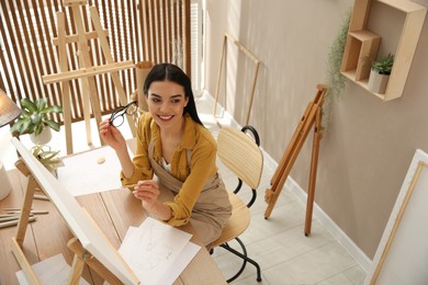 Photo of Young woman drawing on easel with pencil at table indoors, above view