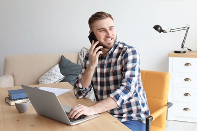 Photo of Young man talking on mobile phone while working with laptop at desk. Home office