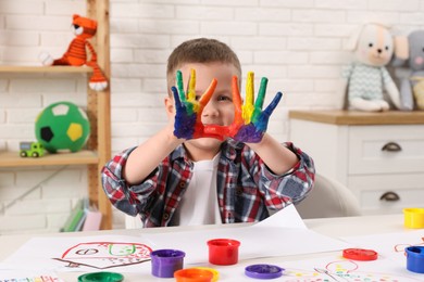 Photo of Happy little boy showing painted palms at white table in room