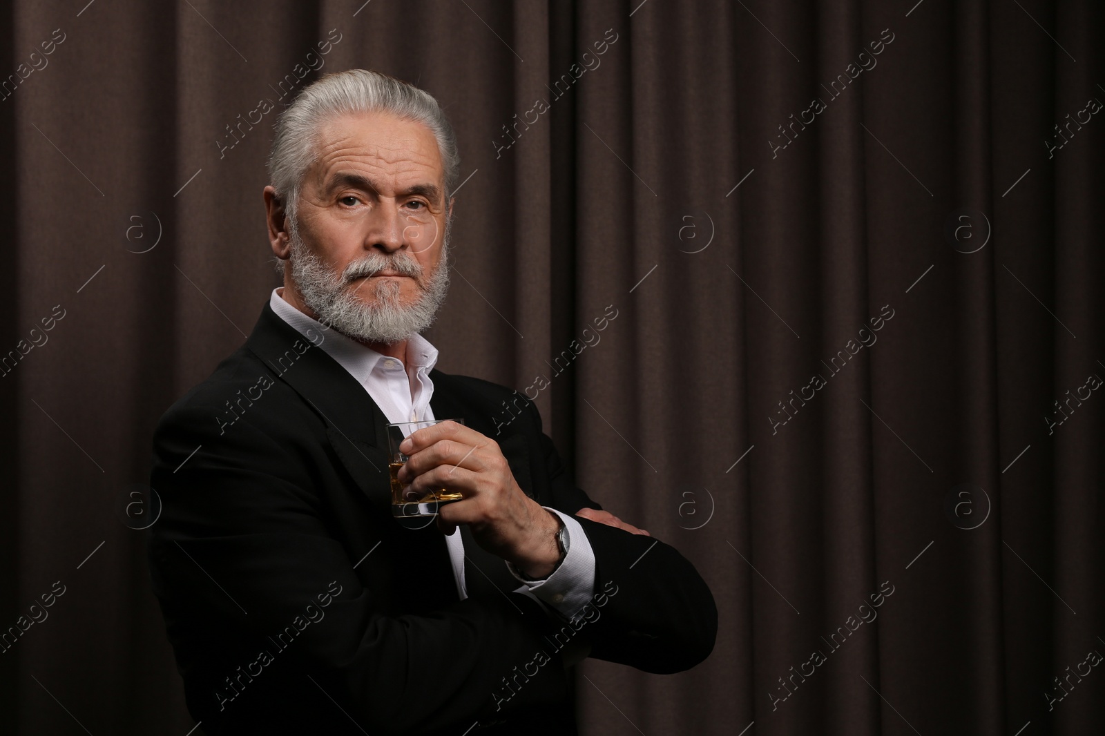 Photo of Senior man in suit holding glass of whiskey with ice cubes on brown background. Space for text