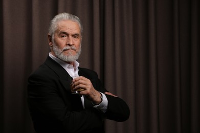 Photo of Senior man in suit holding glass of whiskey with ice cubes on brown background. Space for text