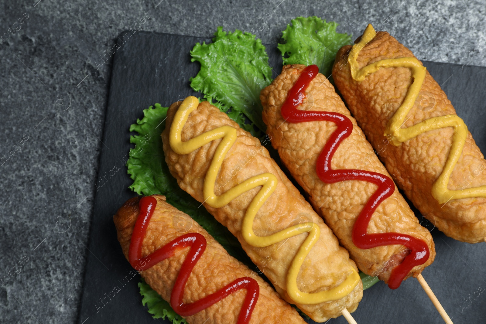 Photo of Delicious deep fried corn dogs with lettuce and sauces on grey table, top view