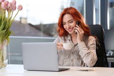 Happy woman with cup of coffee talking on smartphone while working with laptop in office