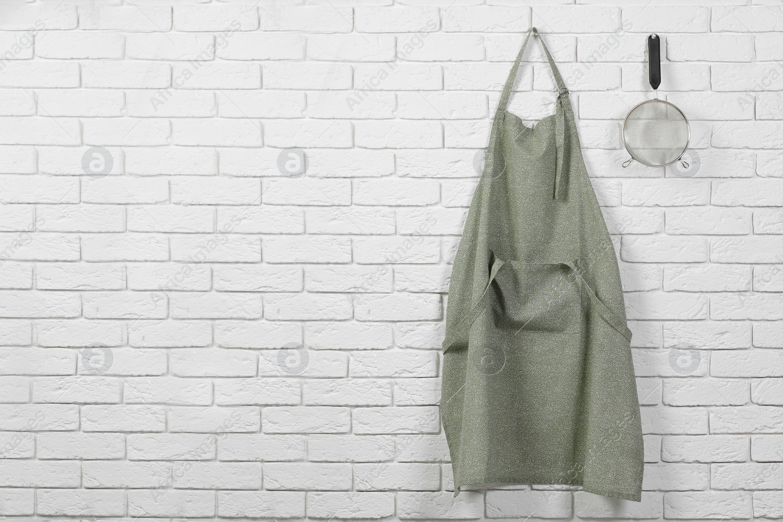 Photo of Clean kitchen apron with pattern and sieve on white brick wall. Space for text