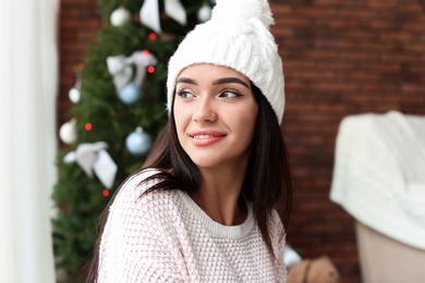 Photo of Beautiful young woman in hat near Christmas tree at home