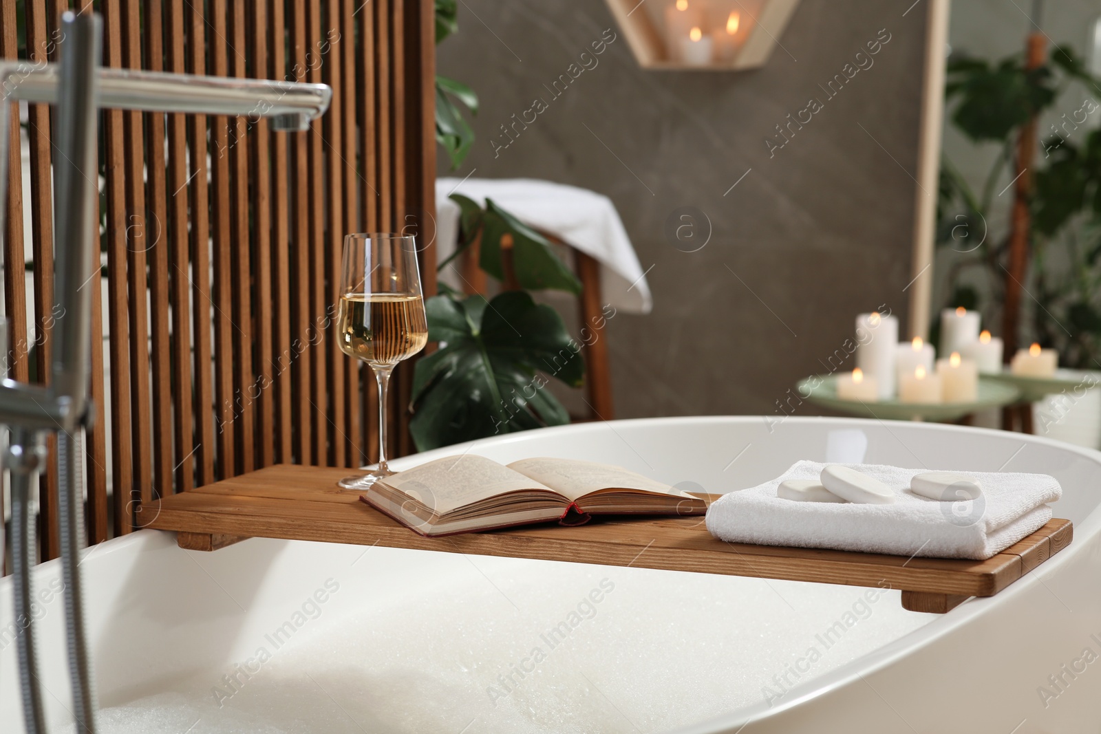 Photo of Wooden bath tray with glass of wine, open book, massage stones and towel on tub indoors. Relaxing atmosphere