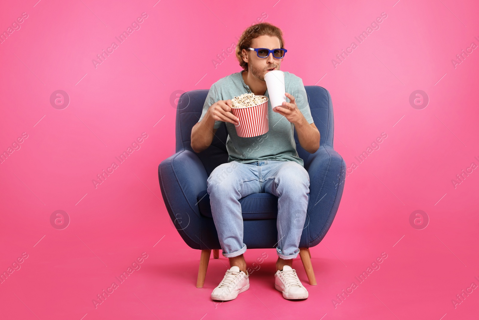 Photo of Emotional man with 3D glasses, popcorn and beverage sitting in armchair during cinema show on color background