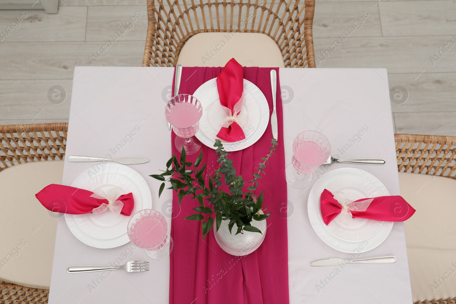 Photo of Table setting. Glasses of tasty beverage, plates, pink napkins and vase with green branches in dining room, above view