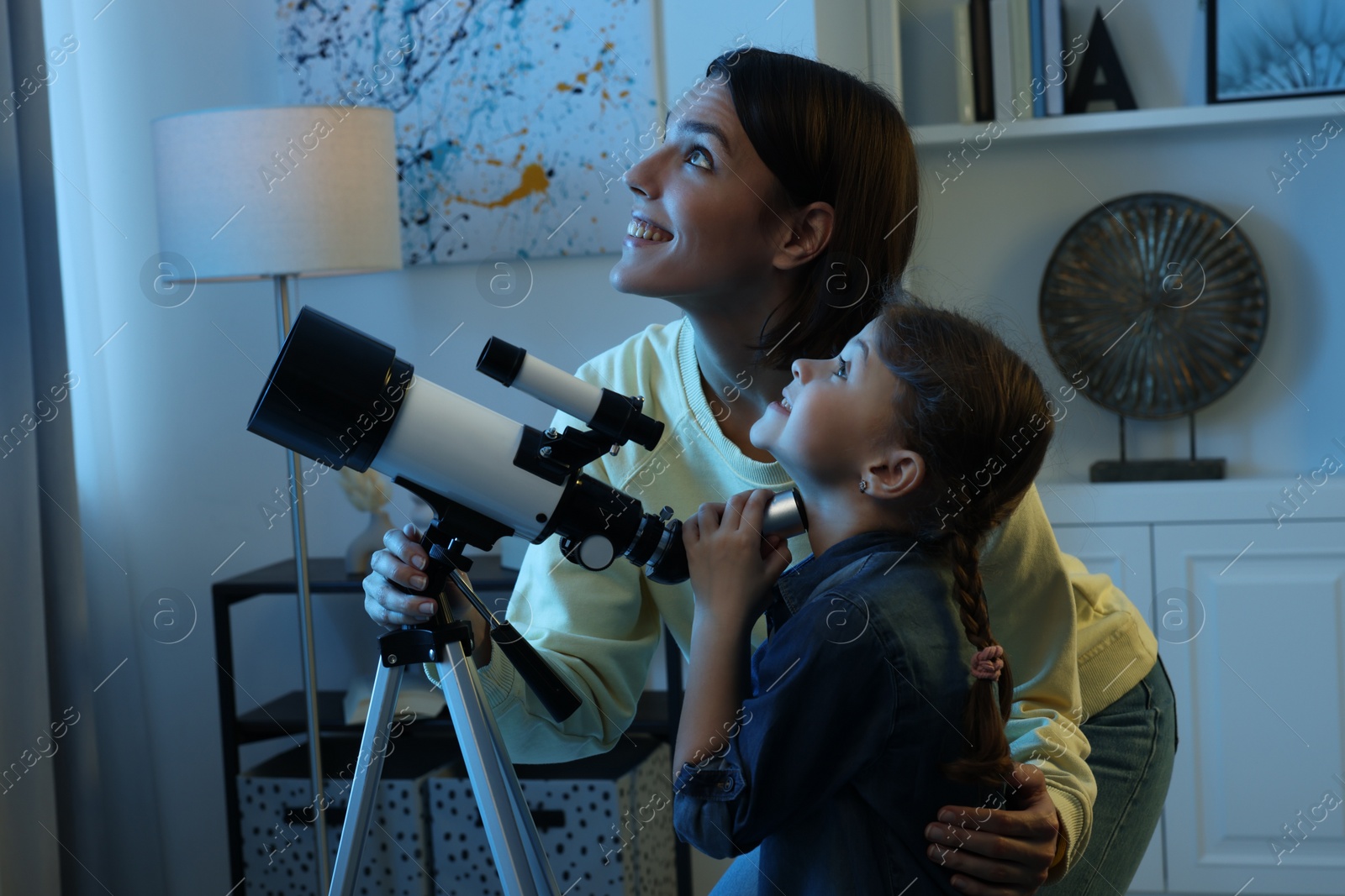 Photo of Happy mother and her cute daughter using telescope to look at stars in room