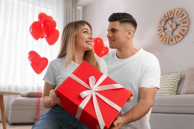 Photo of Man presenting gift to his girlfriend in room decorated with heart shaped balloons. Valentine's day celebration