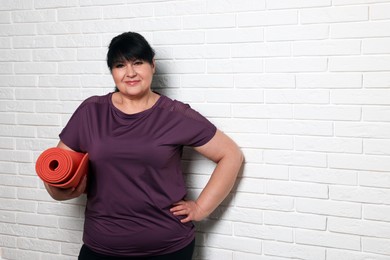 Happy overweight mature woman with yoga mat near white brick wall, space for text