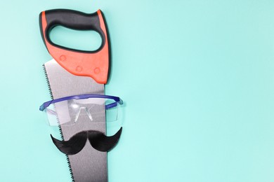 Man's face made of artificial mustache, safety glasses and hand saw on light blue background, top view. Space for text