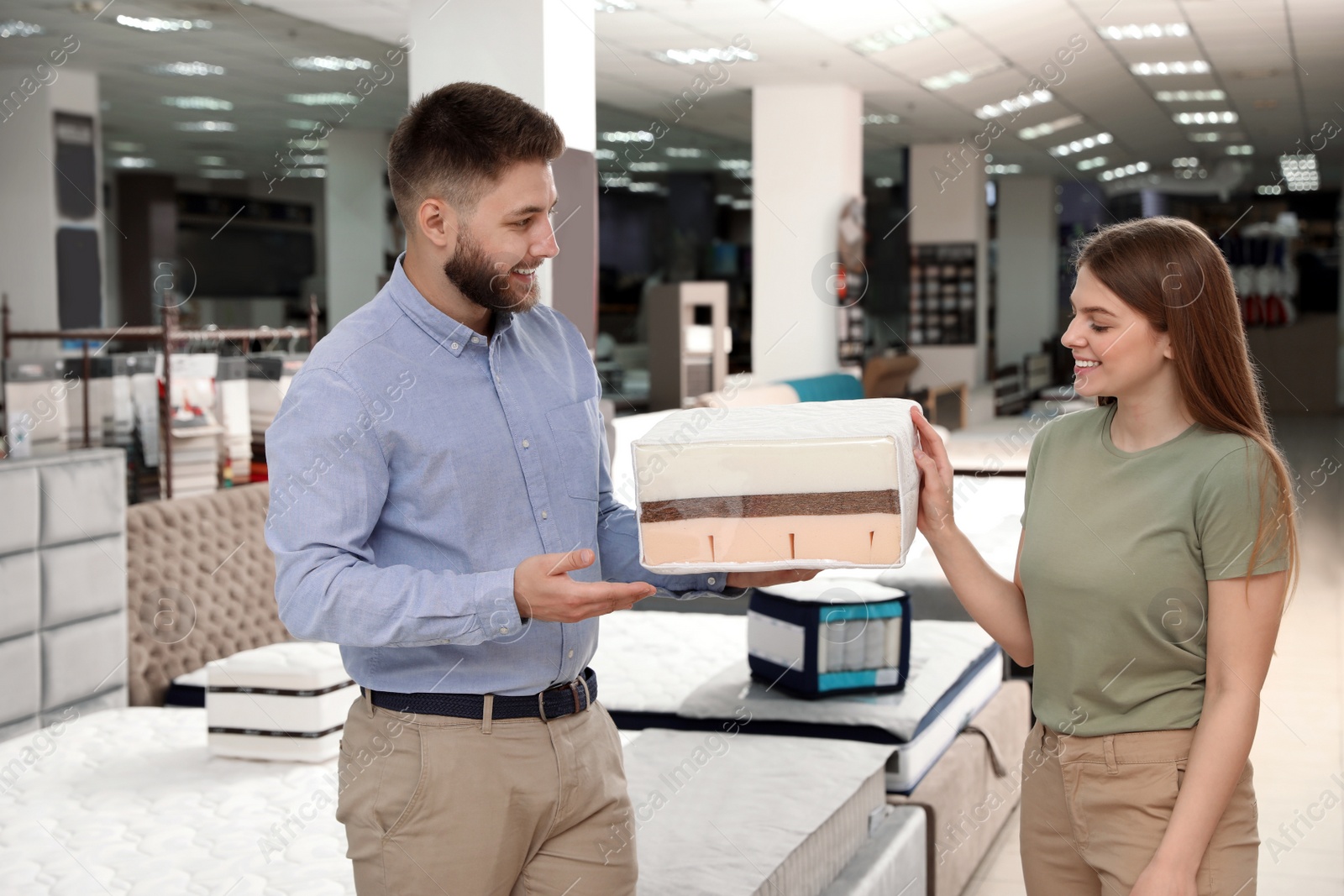 Photo of Young salesman showing sample of modern orthopedic mattress to customer in store