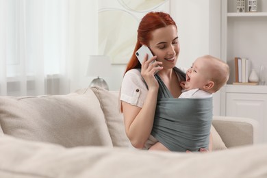 Photo of Mother talking on smartphone while holding her child in sling (baby carrier) at home