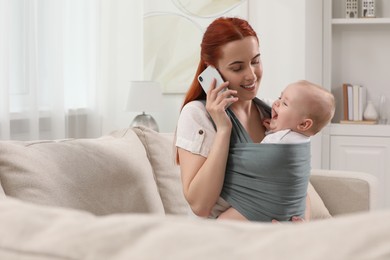 Mother talking on smartphone while holding her child in sling (baby carrier) at home