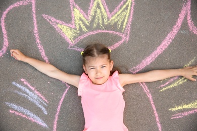 Photo of Little child lying near chalk drawing of wings and crown on asphalt, top view