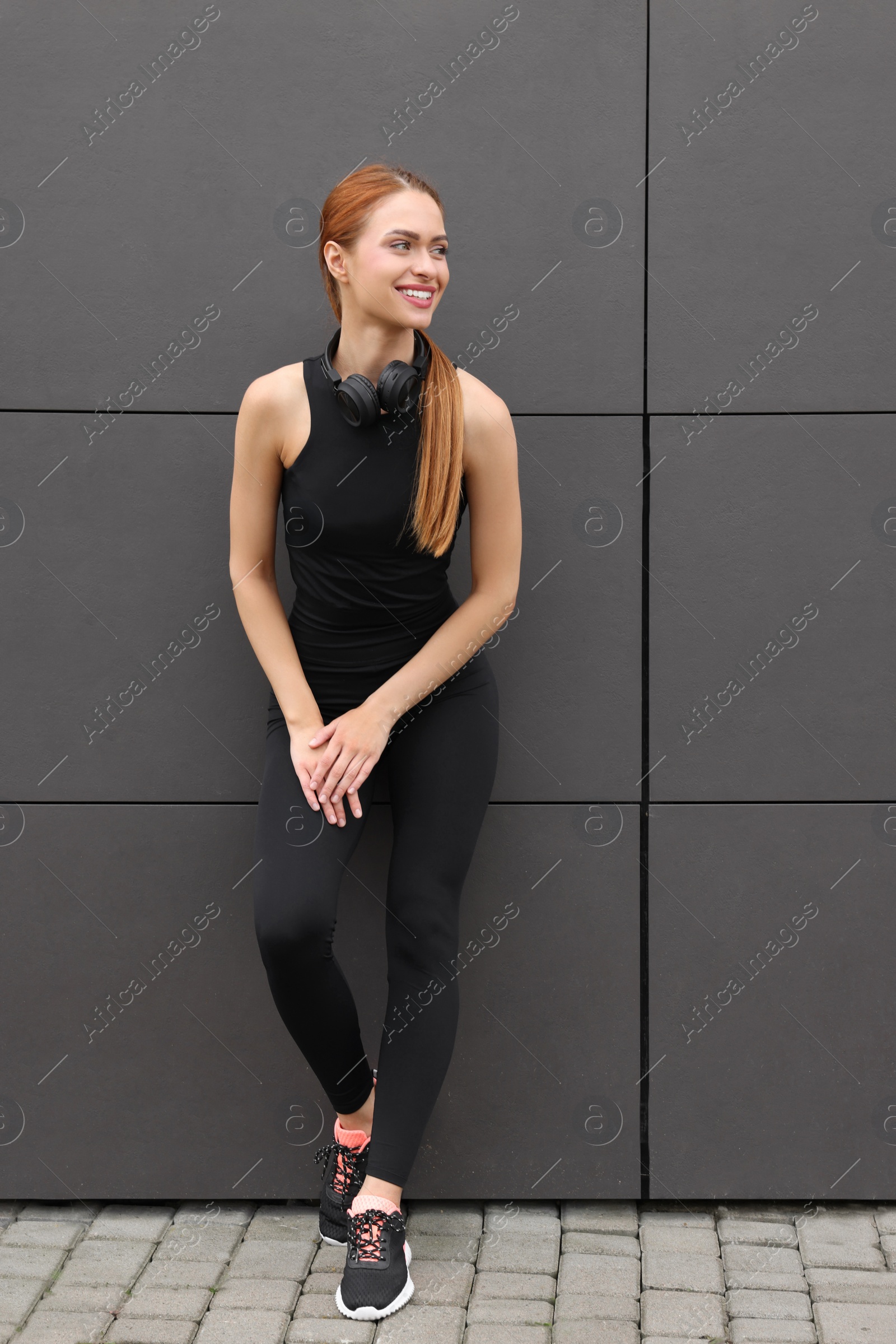 Photo of Beautiful woman in stylish gym clothes with headphones near dark grey wall on street, space for text