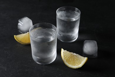 Photo of Shot glasses of vodka with lemon slices and ice on black background