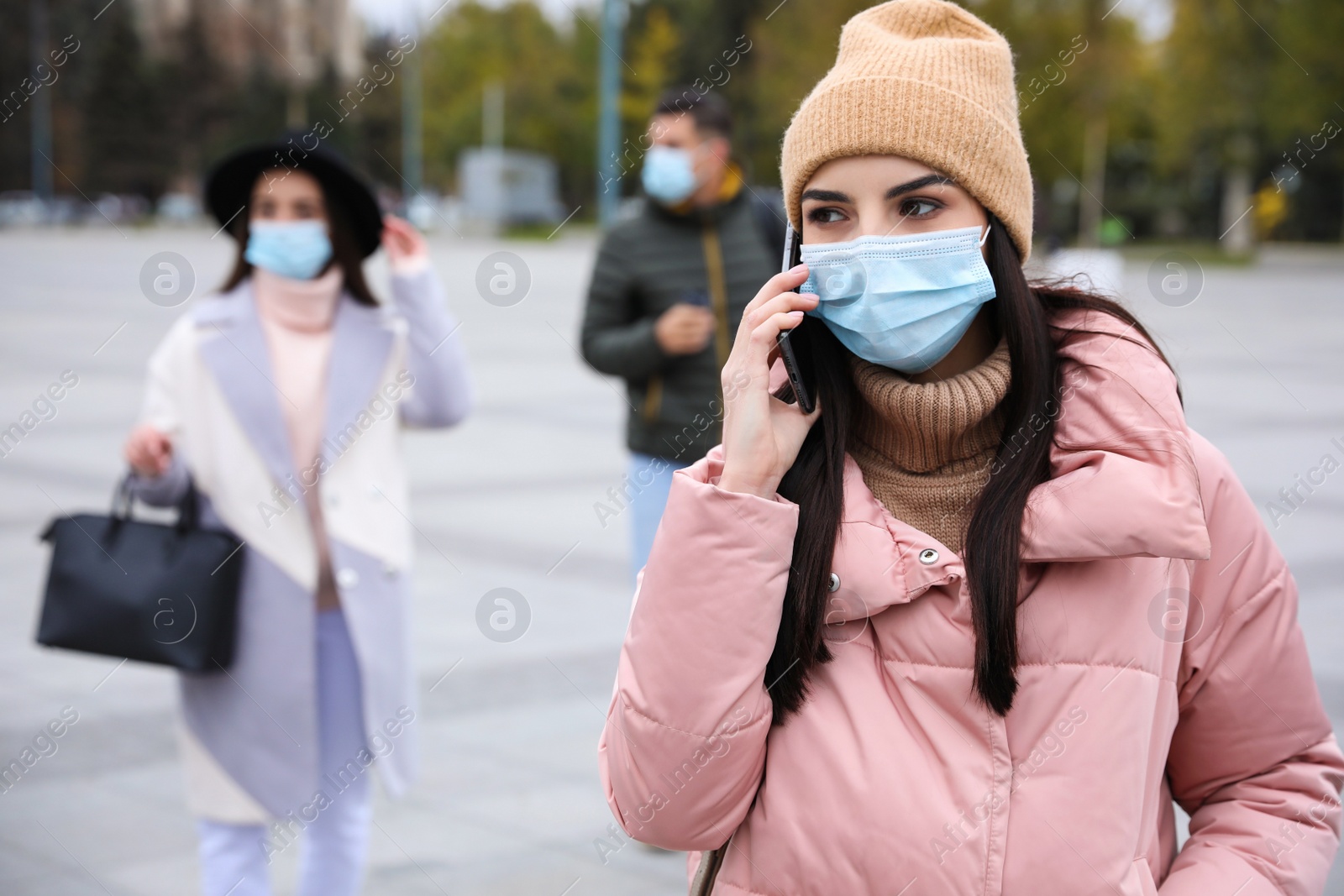 Photo of Young woman in medical face mask talking on phone while walking outdoors. Personal protection during COVID-19 pandemic