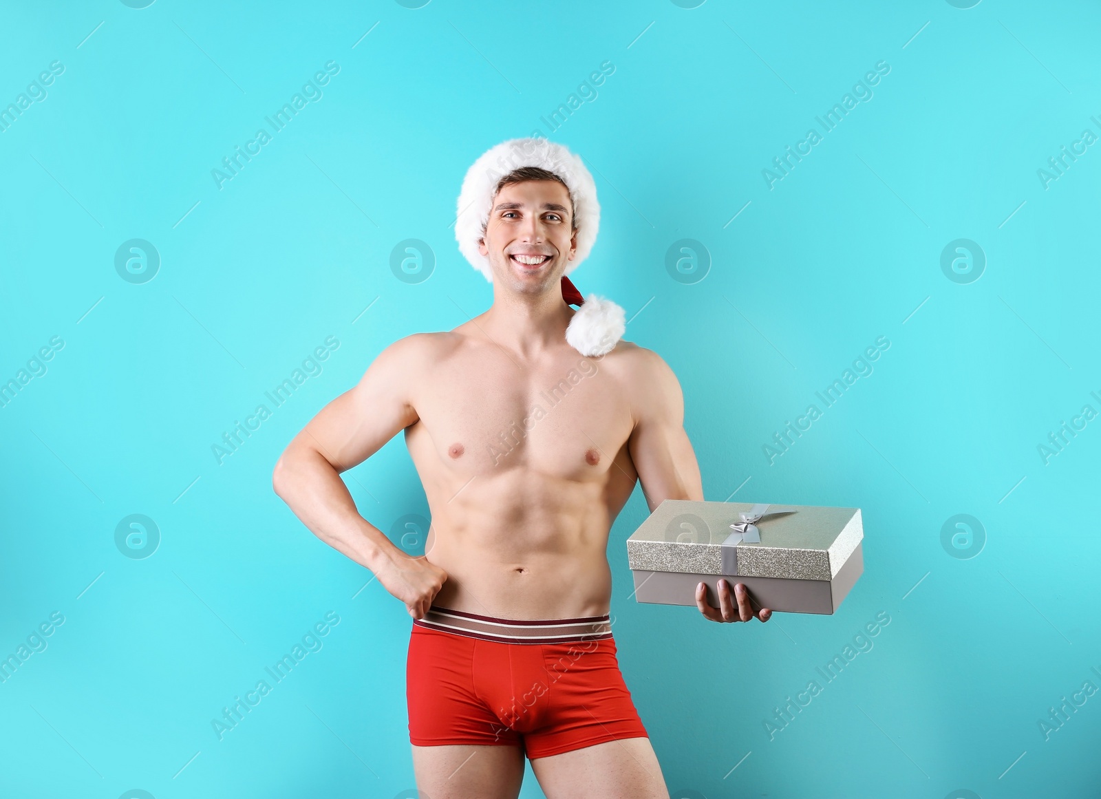 Photo of Young muscular man in Santa hat with gift box on color background