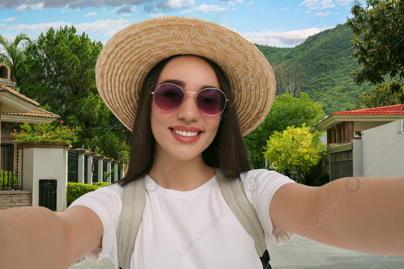 Image of Smiling young woman in sunglasses and straw hat taking selfie in city