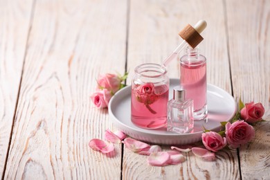 Bottles of essential rose oil and flowers on white wooden table, space for text