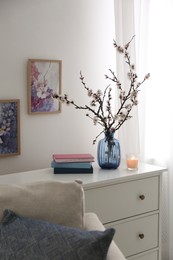 Photo of Flowering tree twigs, burning candle and books on white chest of drawers in living room