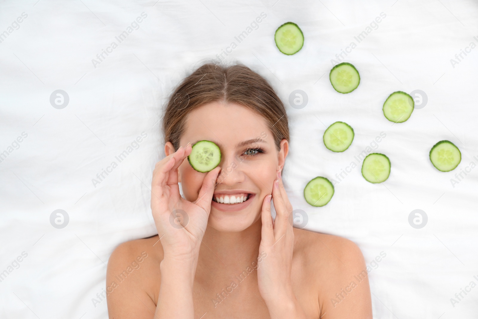 Photo of Beautiful woman with cucumber slices on white fabric, above view. Organic face mask