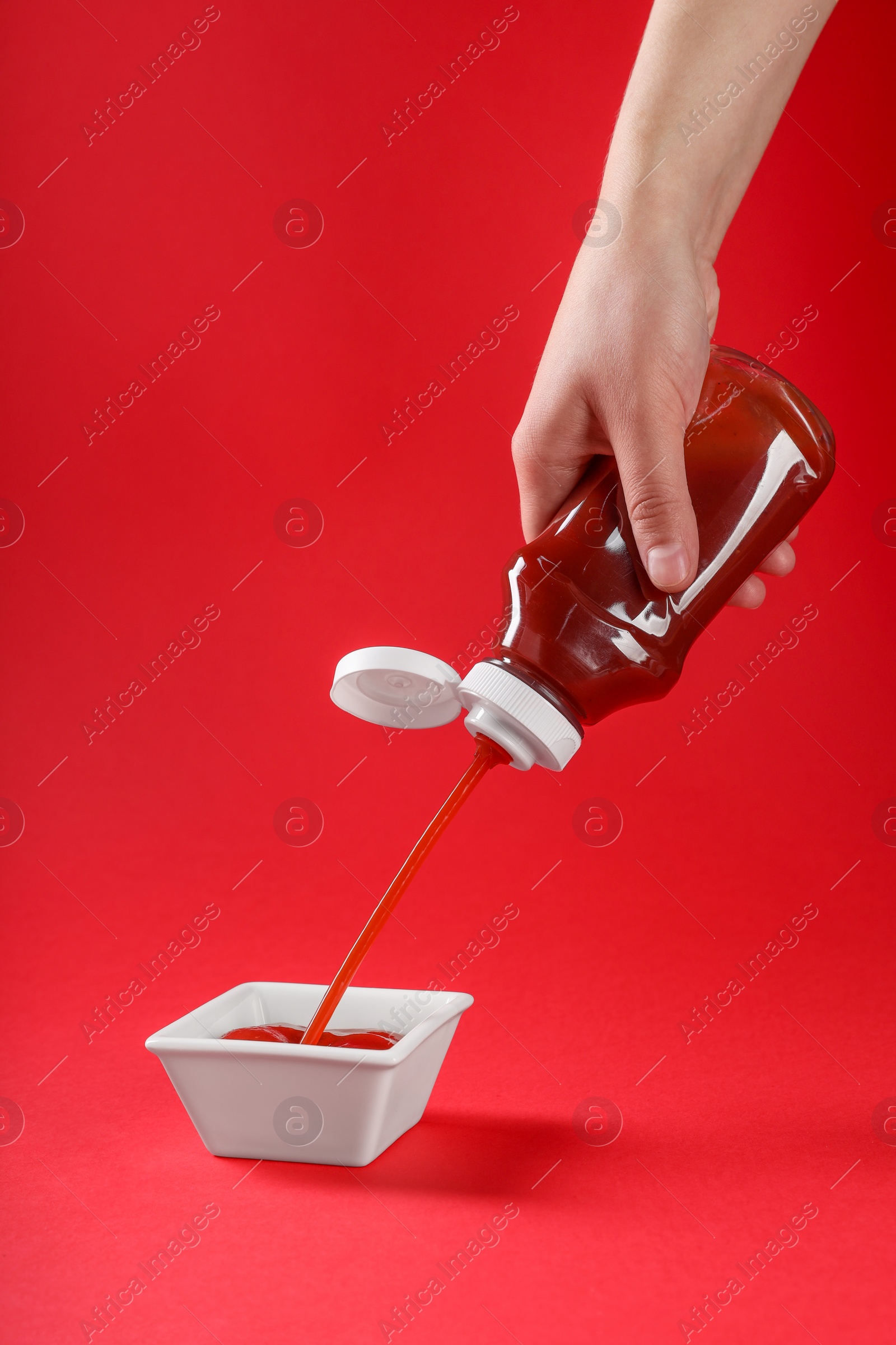 Photo of Woman pouring tasty ketchup from bottle into bowl on red background, closeup