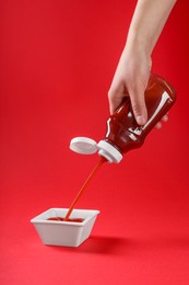 Woman pouring tasty ketchup from bottle into bowl on red background, closeup