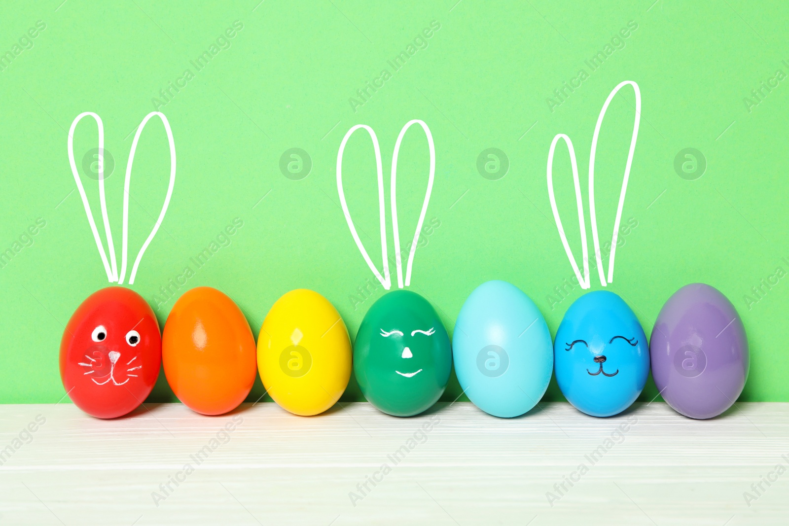 Image of Several eggs with drawn faces and ears as Easter bunnies among others on white wooden table against green background