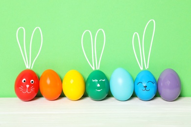 Image of Several eggs with drawn faces and ears as Easter bunnies among others on white wooden table against green background