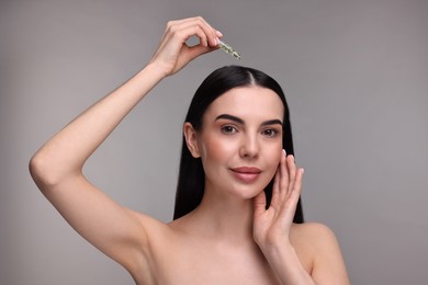 Beautiful young woman using ampoule for hair treatment on grey background