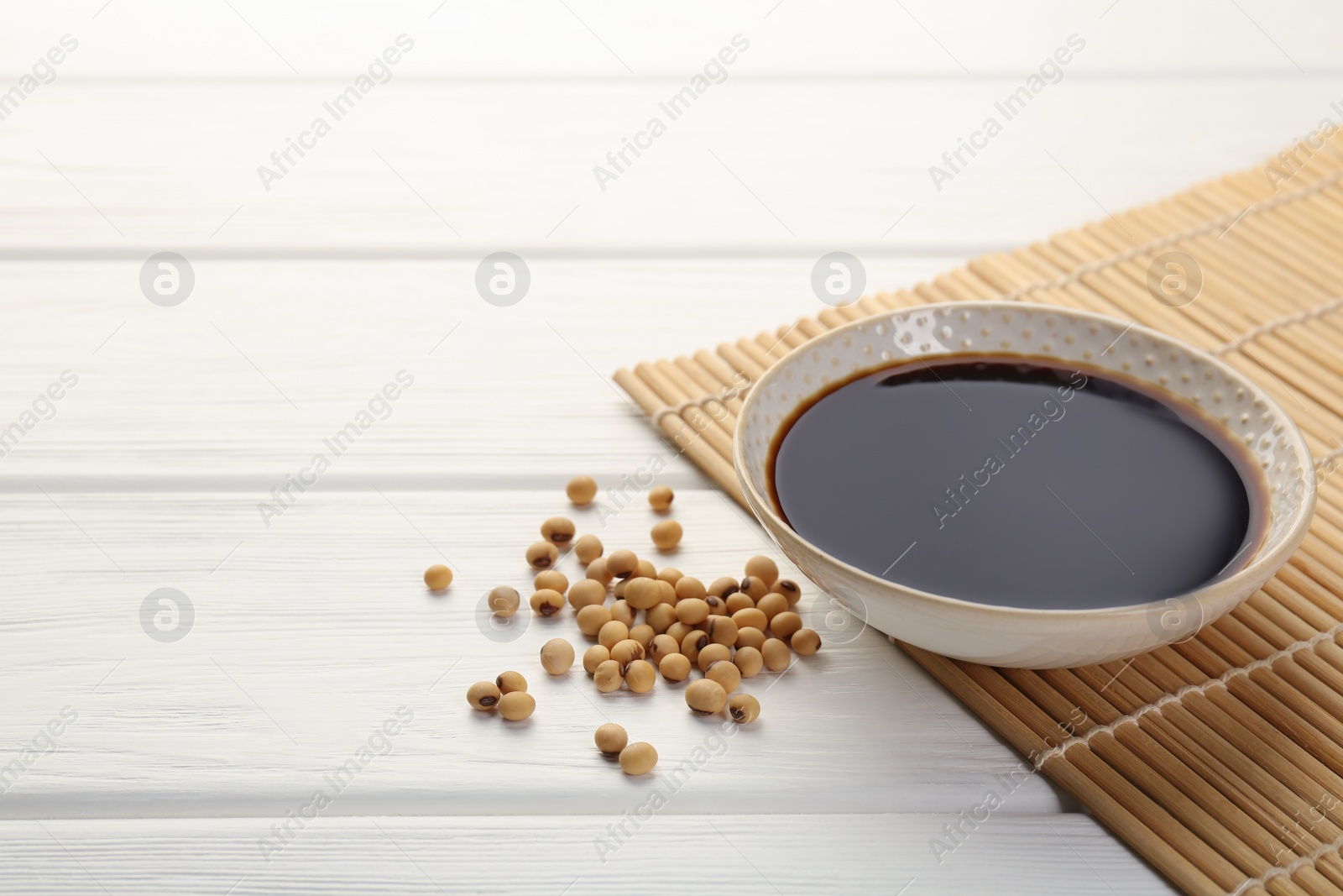 Photo of Tasty soy sauce in bowl and soybeans on white wooden table, closeup. Space for text
