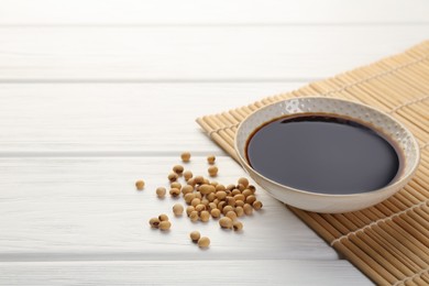 Tasty soy sauce in bowl and soybeans on white wooden table, closeup. Space for text