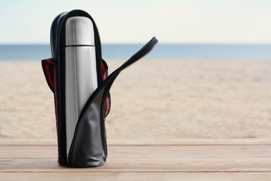 Photo of Metallic thermos in black stylish case on wooden surface near sea, space for text
