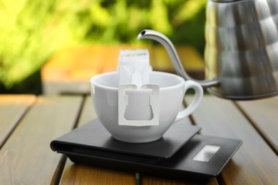 Photo of Cup with drip coffee bag, kettle and kitchen scales on wooden table, closeup