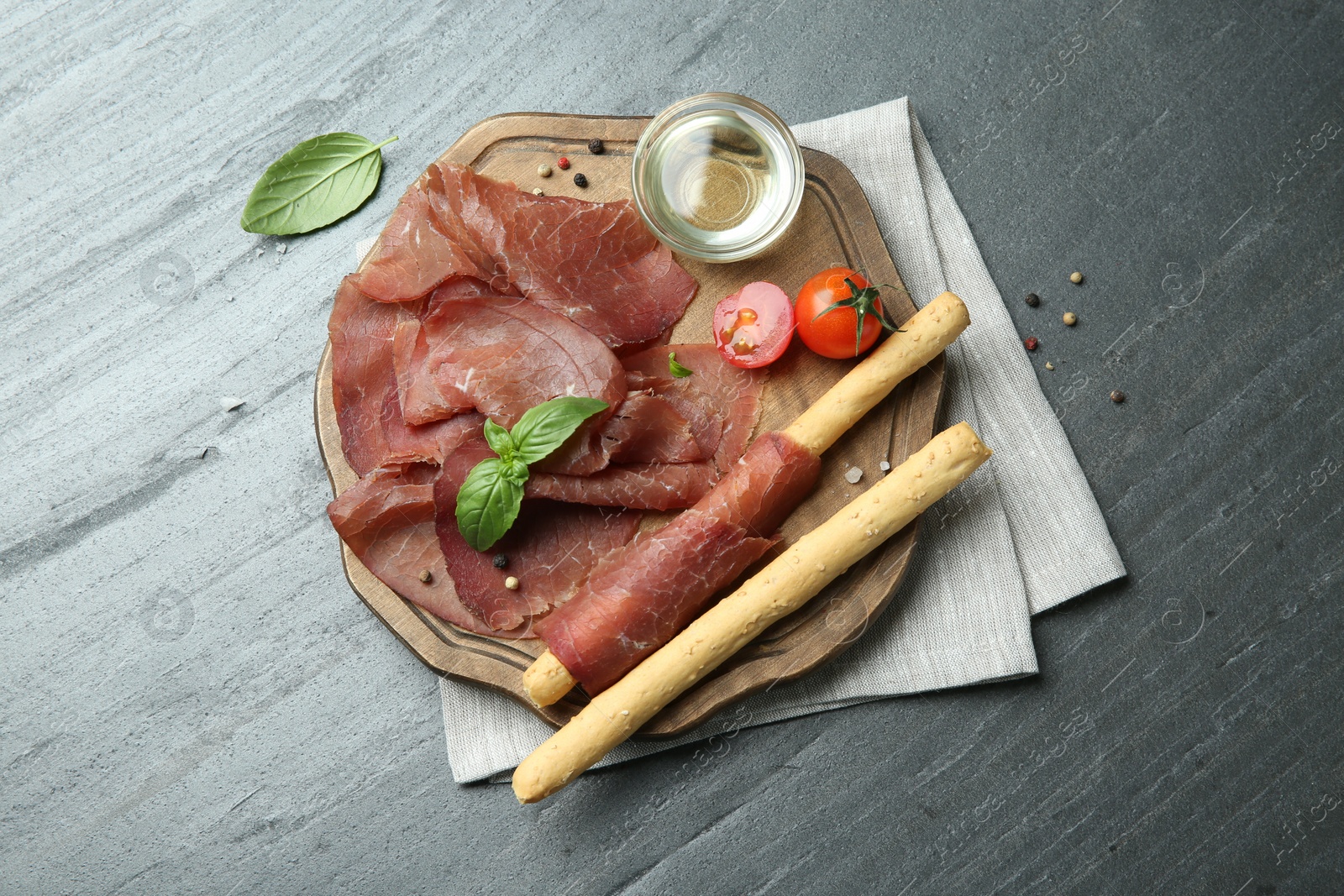 Photo of Delicious bresaola, tomato, grissini sticks and basil leaves on grey textured table, top view